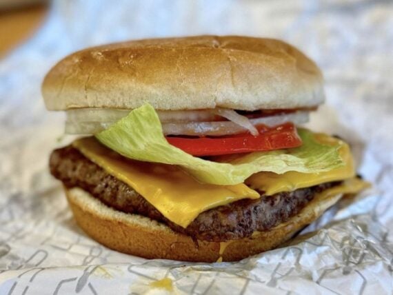 A cheeseburger with lettuce, tomato, onion, and two slices of cheddar cheese on a beef patty in a soft bun, resting on crumpled white paper.