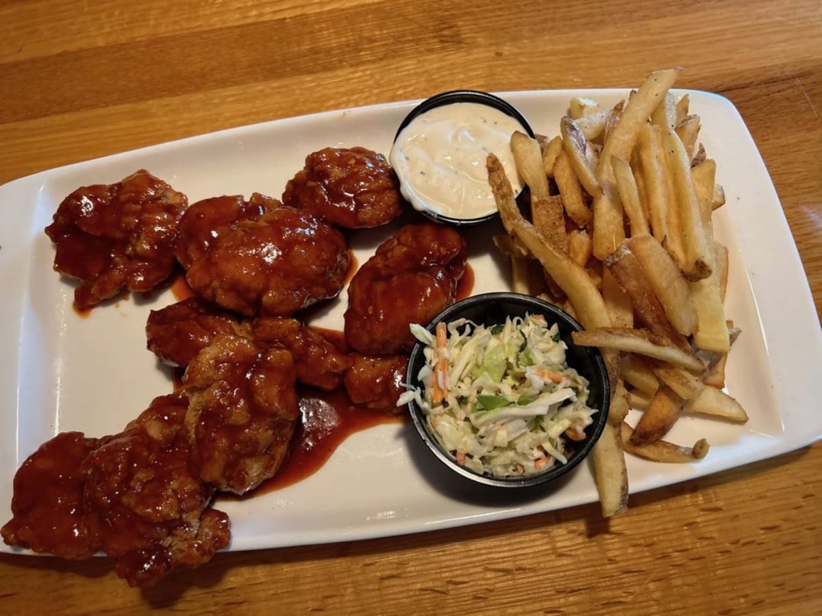 A rectangular white plate with boneless chicken wings covered in sauce, French fries, ranch dressing, and coleslaw on a wooden table—perfect for enjoying all-you-can-eat deals under $20.
