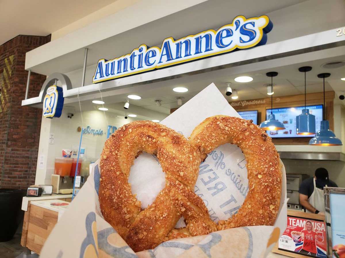 A hand holds a salted pretzel in front of an Auntie Anne’s store counter, with the bright blue and yellow Auntie Anne’s sign visible above the counter in the background.