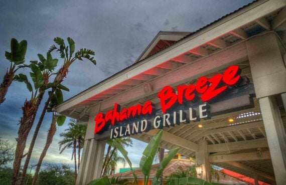 A Bahama Breeze Island Grille restaurant entrance features a bold red neon sign under a peaked roof, surrounded by lush palm trees and tropical plants, capturing the laid-back spirit of Bahama Breeze against a cloudy sky.