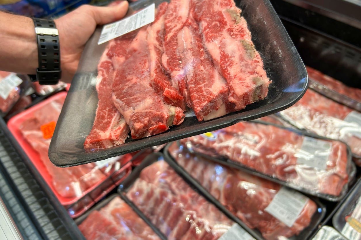 A hand holds a packaged tray of raw beef ribs with a price label, above a refrigerated display filled with similar packages of meat at a grocery store.