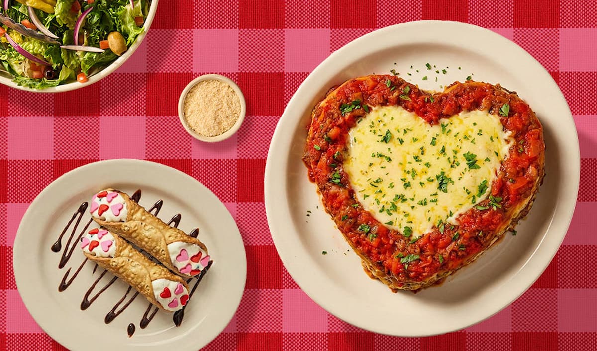 A heart-shaped baked pasta dish with tomato sauce and melted cheese sits on a plate, next to two decorated cannoli on a separate plate, a small bowl of grated cheese, and a salad, all on a red checkered tablecloth.