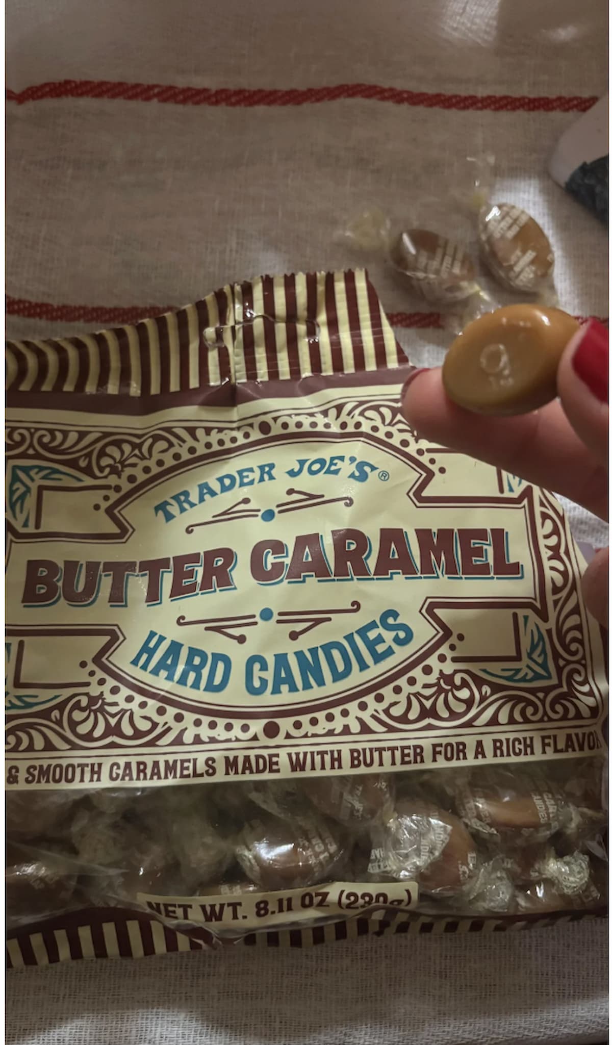 A hand holds a wrapped butter caramel candy in front of an open bag labeled "Trader Joe’s Butter Caramel Hard Candies,” with more candies visible in the bag and on the table.