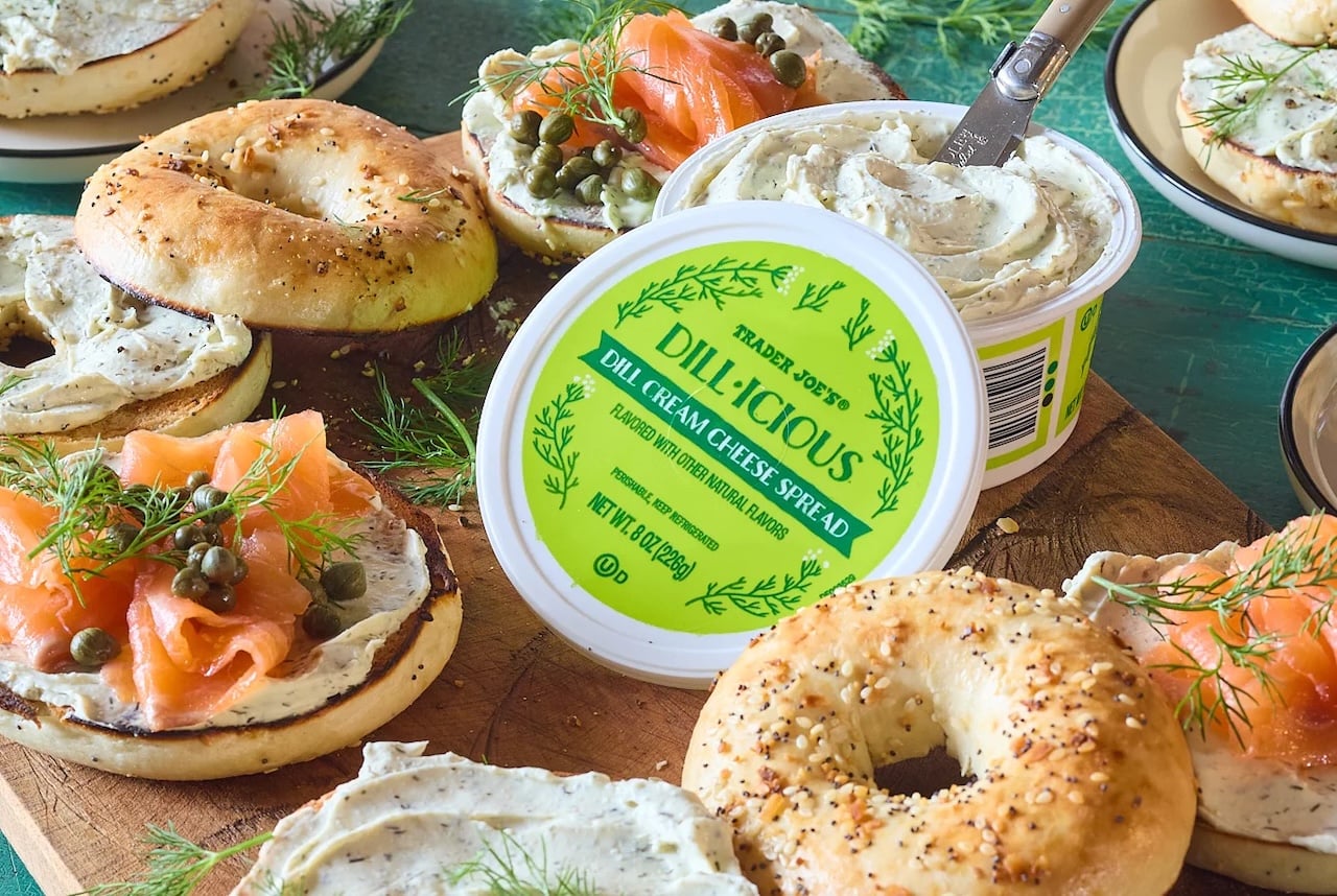 A container of Trader Joe’s Dill-icious Dill Cream Cheese Spread is open on a wooden board, surrounded by bagels topped with the spread, smoked salmon, capers, and dill. Some bagels are plain; others have seeds.
