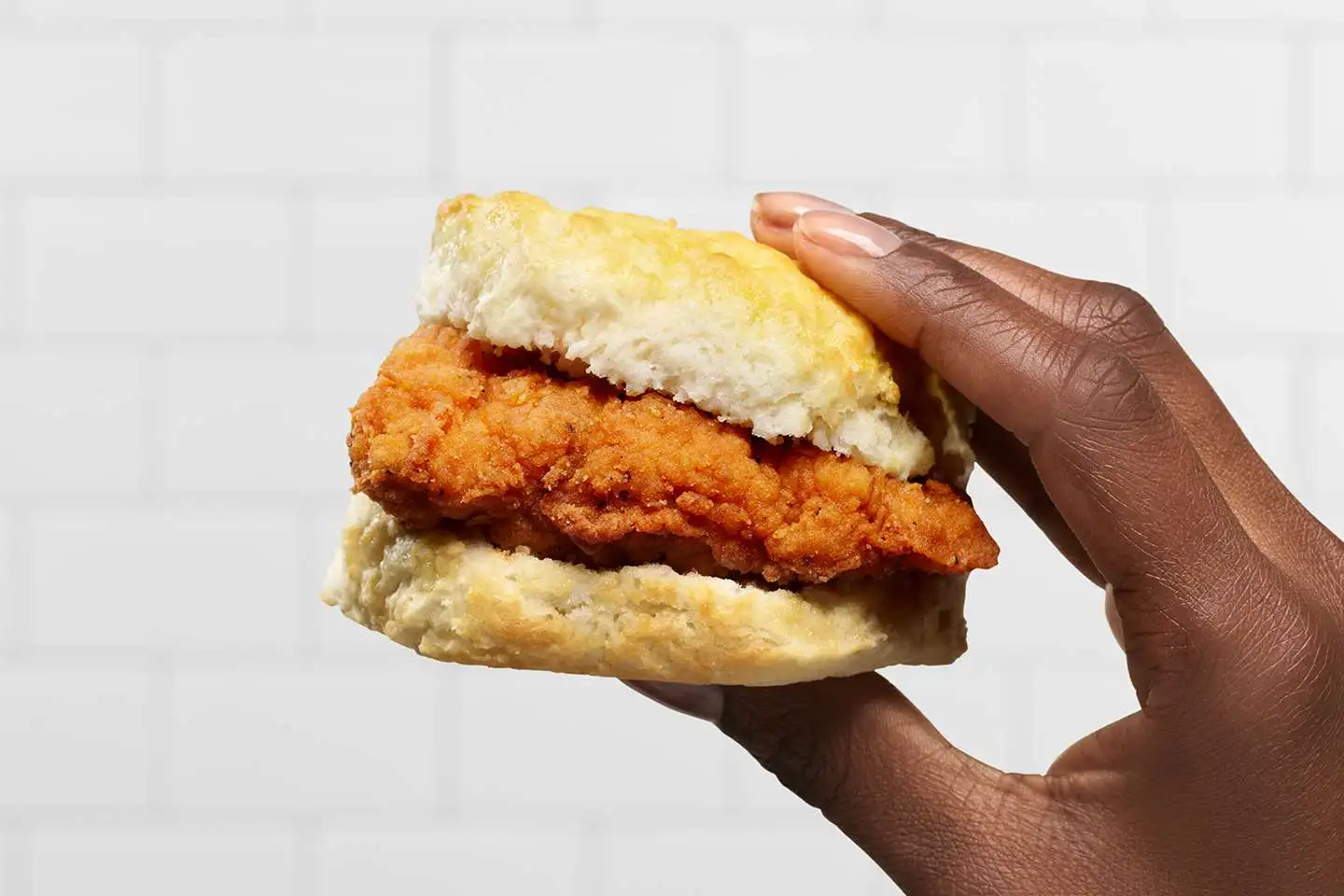 A hand holds a biscuit sandwich filled with a piece of crispy fried chicken against a white tiled background.