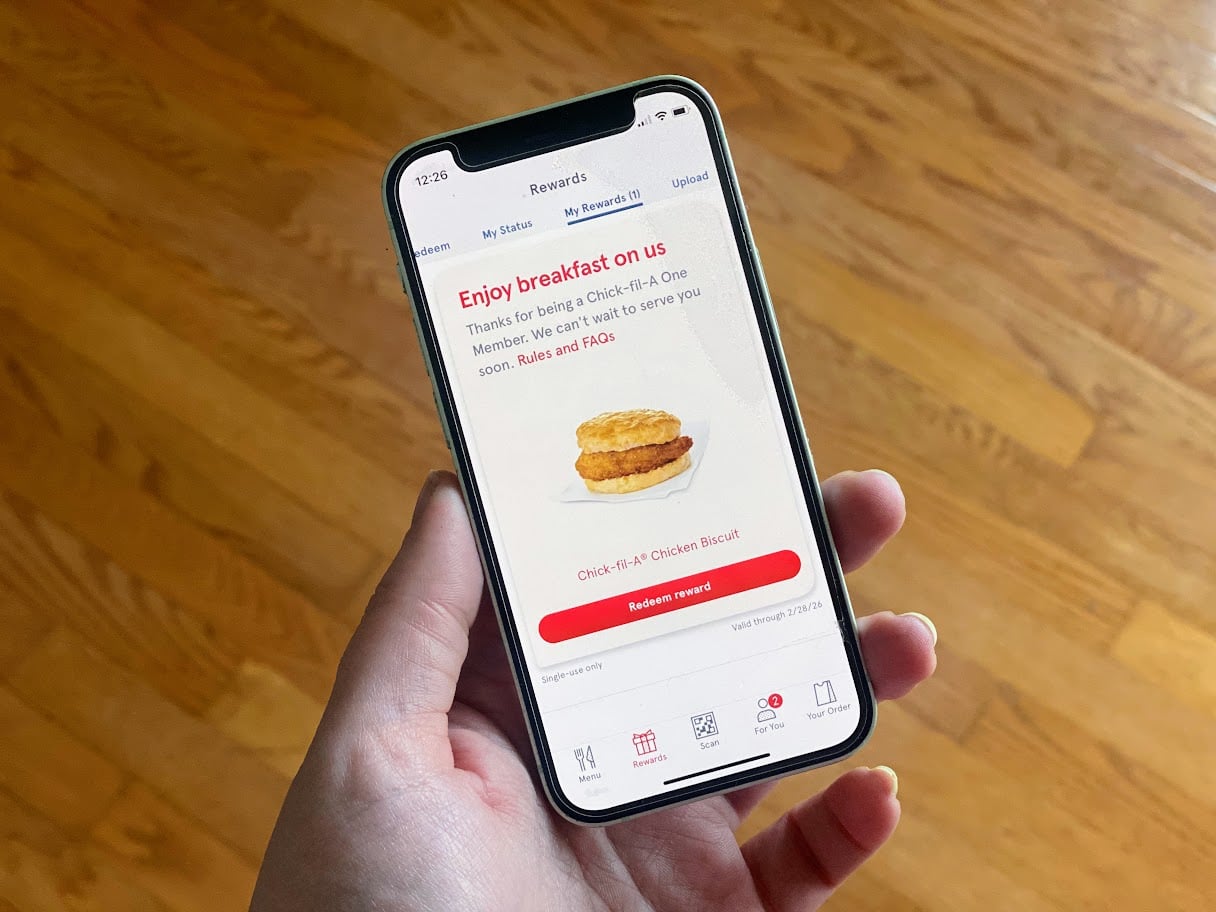 A hand holds a smartphone displaying a Chick-fil-A rewards screen offering a free Chick-fil-A chicken biscuit. The phone is against a wooden floor background.