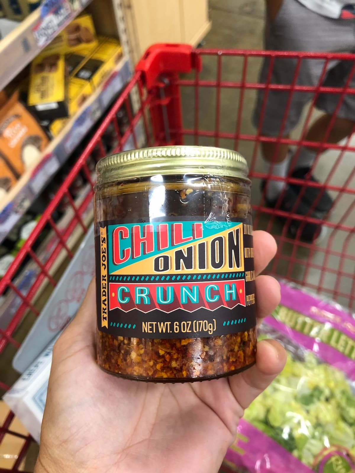 A hand holds a jar of Trader Joe’s Chili Onion Crunch in a shopping cart, surrounded by grocery shelves—proof that even if you think Trader Joe’s is overrated, this popular condiment still stands out.