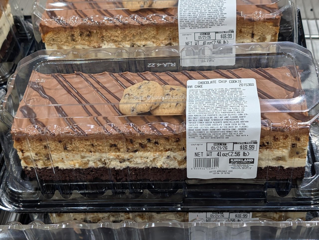 A rectangular chocolate chip cookie cake in a clear plastic container, topped with chocolate drizzle and cookies. The label shows it’s from Kirkland, weighs 4.25 lbs, and is priced at $18.99.