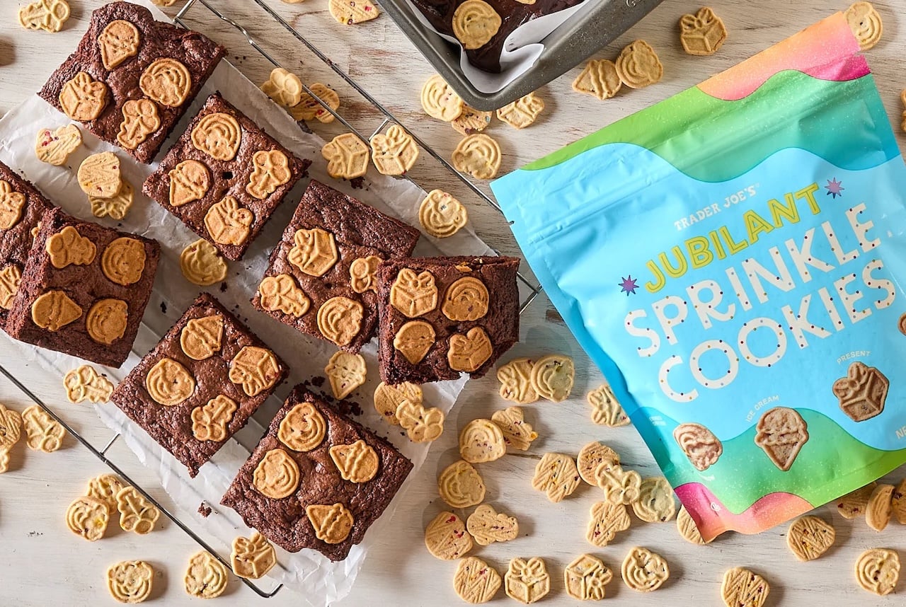 A cooling rack holds brownies topped with small, animal-shaped cookies. More cookies are scattered around, along with a blue and green bag labeled "Trader Joe's Jubilant Sprinkle Cookies.