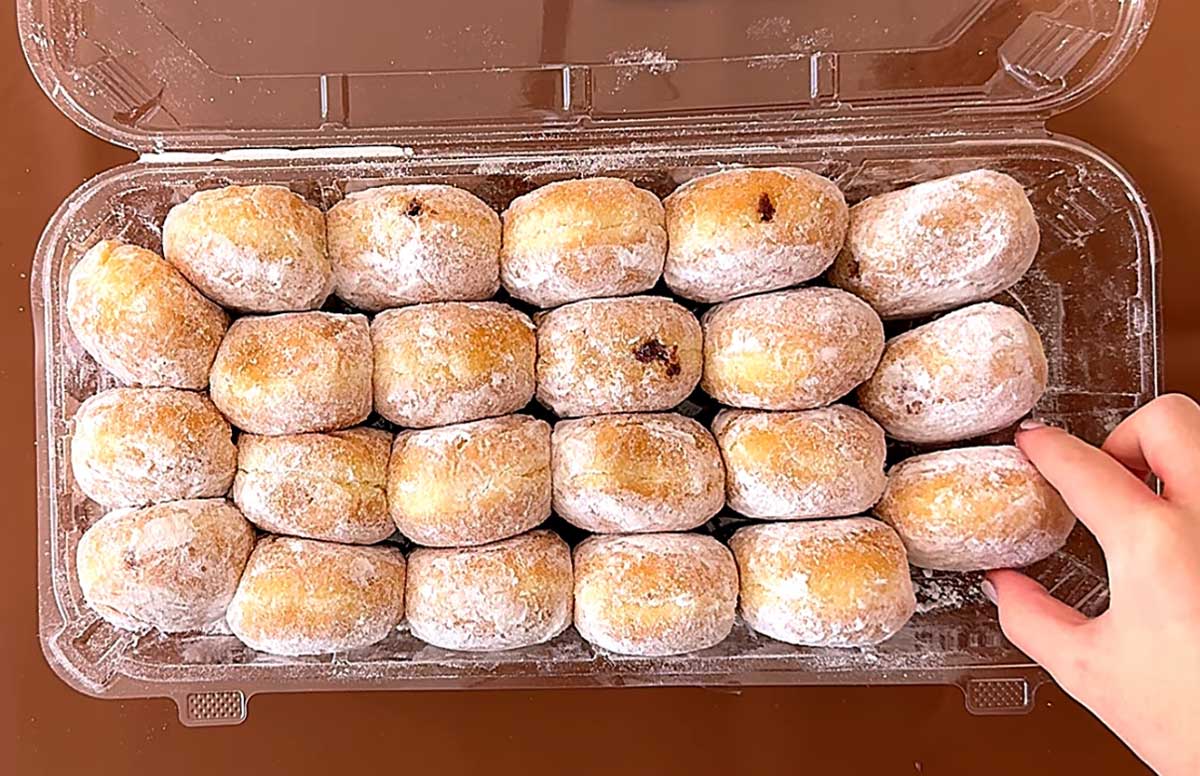 A plastic container holds 24 powdered sugar-coated round doughnuts. A hand on the right side is reaching to pick up one of the doughnuts.