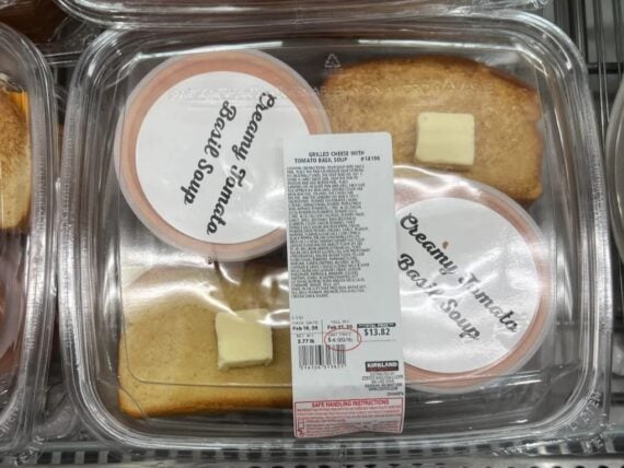 Plastic container with two slices of bread, two pats of butter, and two cups labeled &ldquo;Creamy Tomato Basil Soup.&rdquo; The container has a white price label and is displayed in a refrigerated section.