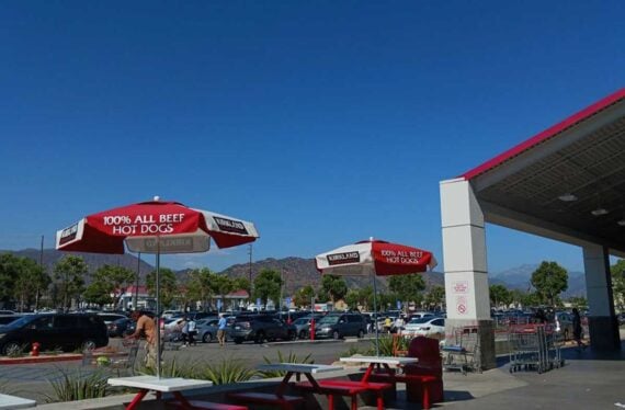 Outdoor seating area with red benches and umbrellas labeled "100% All Beef Hot Dogs" near a parking lot. Cars are parked in the background with mountains and a clear blue sky visible.