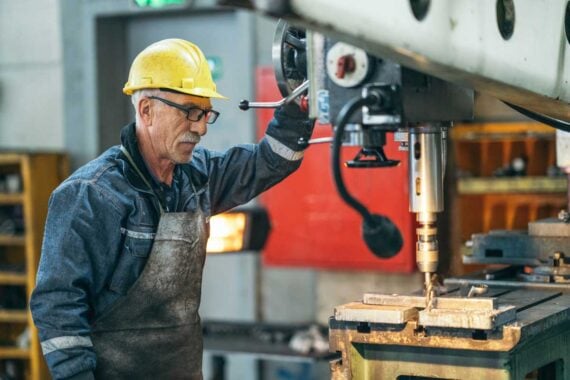 An older man wearing a yellow hard hat, safety glasses, and a denim work shirt operates a large industrial drilling machine in a workshop, representing skilled labor impacted by SNAP work requirements.