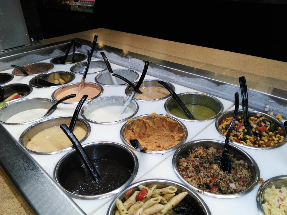 A salad bar with metal bowls containing various dressings, salsas, dips, pasta, and salads—each with a serving spoon—displayed on a refrigerated counter featuring all-you-can-eat deals under $20.