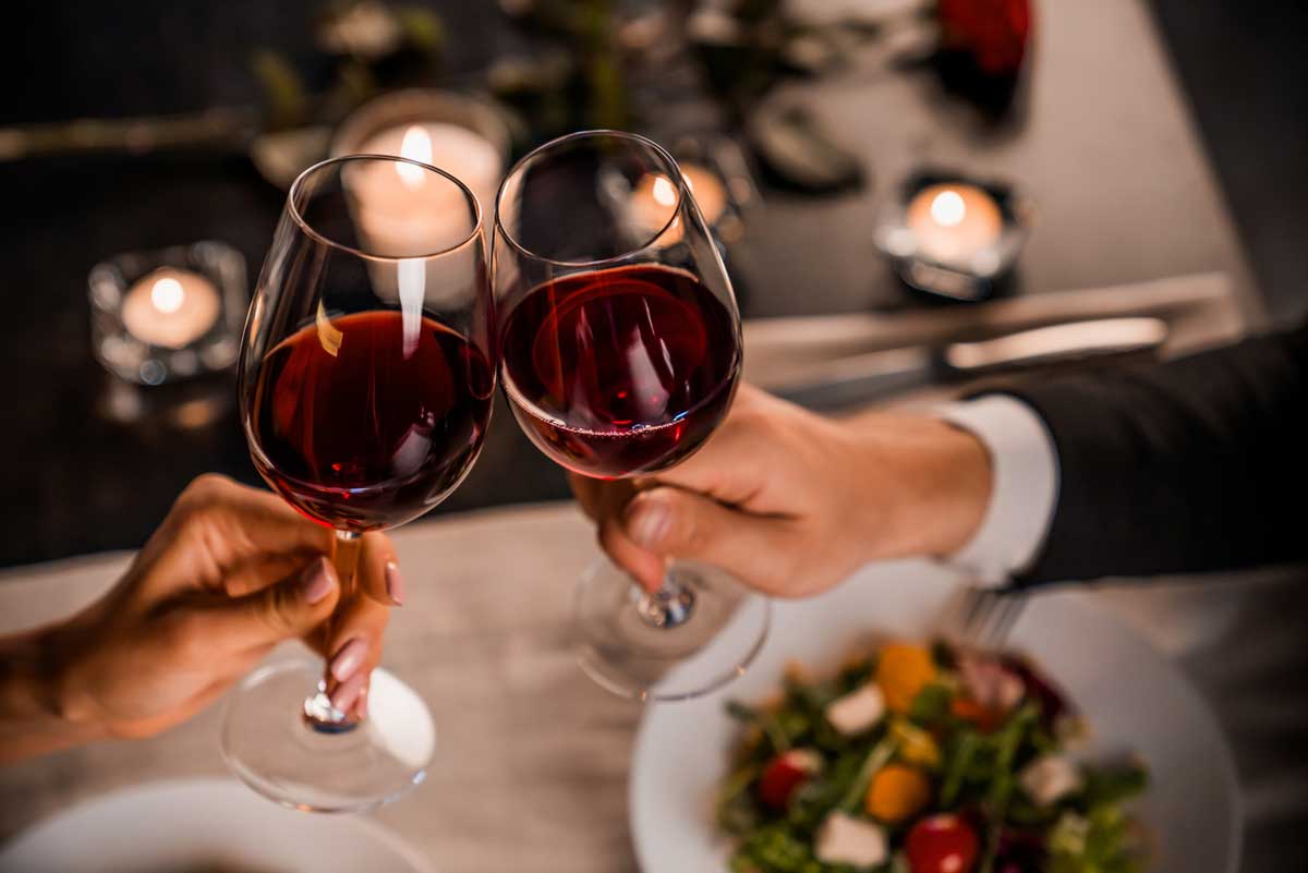 Two people clinking glasses of red wine over a dinner table with a salad, candles, and a romantic atmosphere.