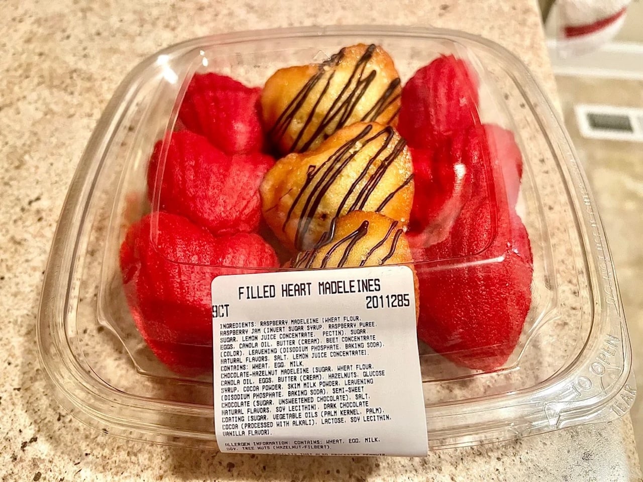 A plastic container holds several red heart-shaped madeleines and two chocolate-drizzled madeleines. A label on the container reads "Filled Heart Madeleines" and lists the ingredients.