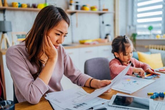 A woman looks stressed while reviewing papers at a table, surrounded by documents, as a young child sits nearby coloring. The homey scene hints at challenges faced in financially distressed cities, with shelves and kitchen items in the background.