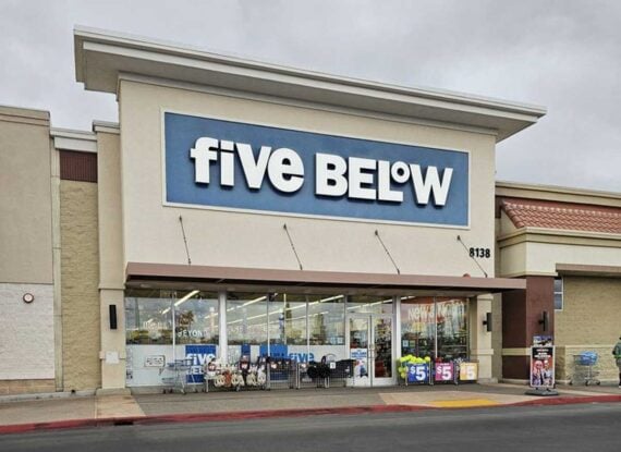 The exterior of a Five Below store with a large blue and white sign above the entrance. Storefront windows display merchandise and bright price signs. The sky is overcast.
