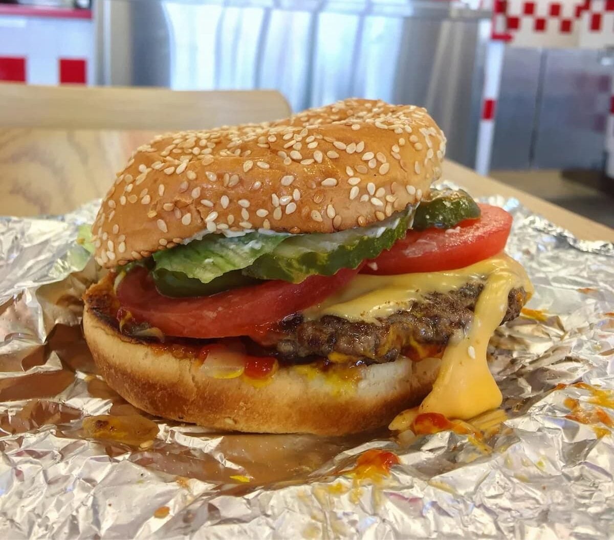 An overrated fast-food burger with lettuce, tomato, pickles, and melted cheese sits on a sheet of foil in a restaurant, with milkshake signs visible in the background.