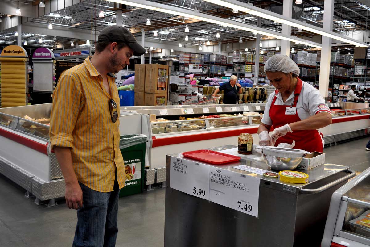 A man in a yellow shirt stands by a food sample station at a grocery store, possibly drawn in by clever grocery store tricks, while a worker in a red apron and hairnet prepares samples. Shelves and freezers are visible in the background.