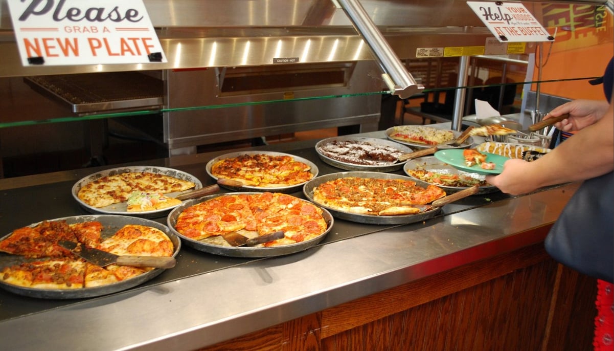 A buffet with various whole pizzas, desserts, and other foods on display. A person serves themselves a slice of pizza onto a green plate. Signs above the counter advertise all-you-can-eat deals under $20 and remind guests to grab a new plate.
