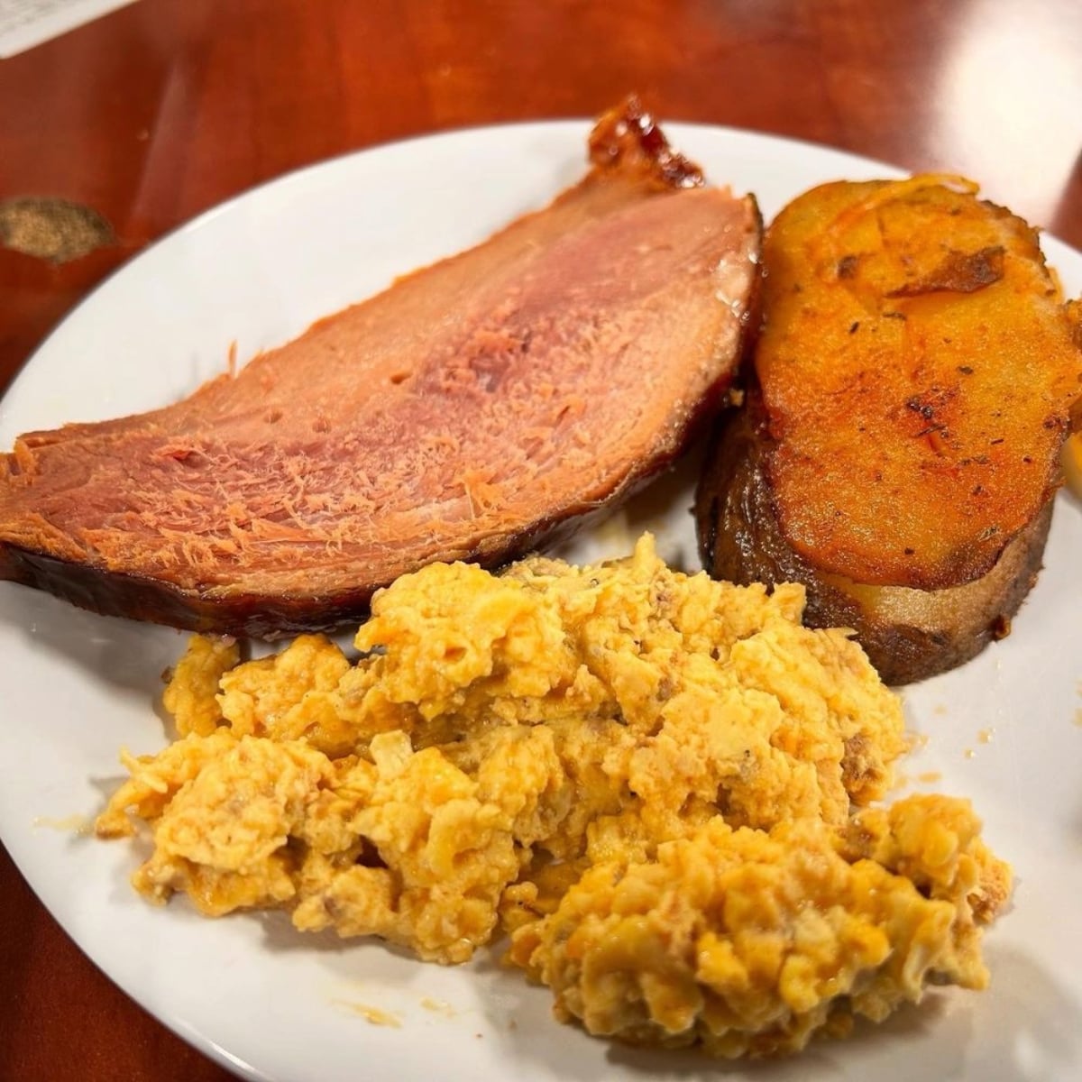 A plate with a slice of baked ham, seasoned baked sweet potato, and scrambled eggs on a wooden table—perfect for those craving hearty all-you-can-eat deals under $20.