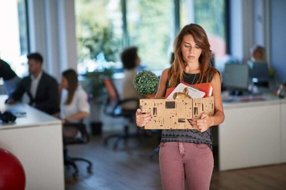 A woman stands in an office holding a box with personal items, looking down sadly, perhaps reflecting on mistakes after losing your job, while other people work in the background at their desks.