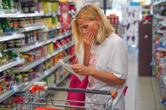 A woman stands in a grocery store aisle, looking worried as she examines a receipt. One hand covers her mouth, and a shopping cart with groceries is in front of her. Shelves of products line both sides.