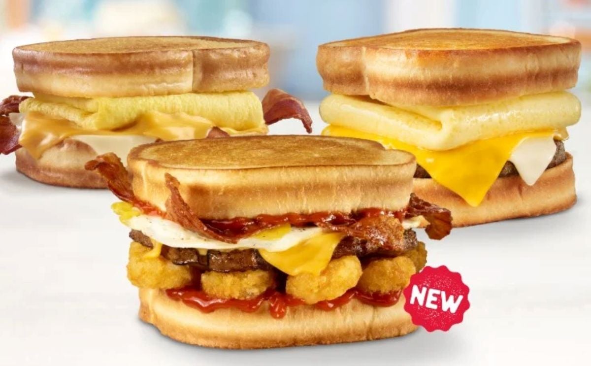 Three breakfast sandwiches on toasted bread are shown from a new fast-food menu. The front sandwich, labeled "NEW," features bacon, egg, cheese, sausage, hashbrowns, and ketchup. The two in the back have eggs, bacon, and melted cheese.
