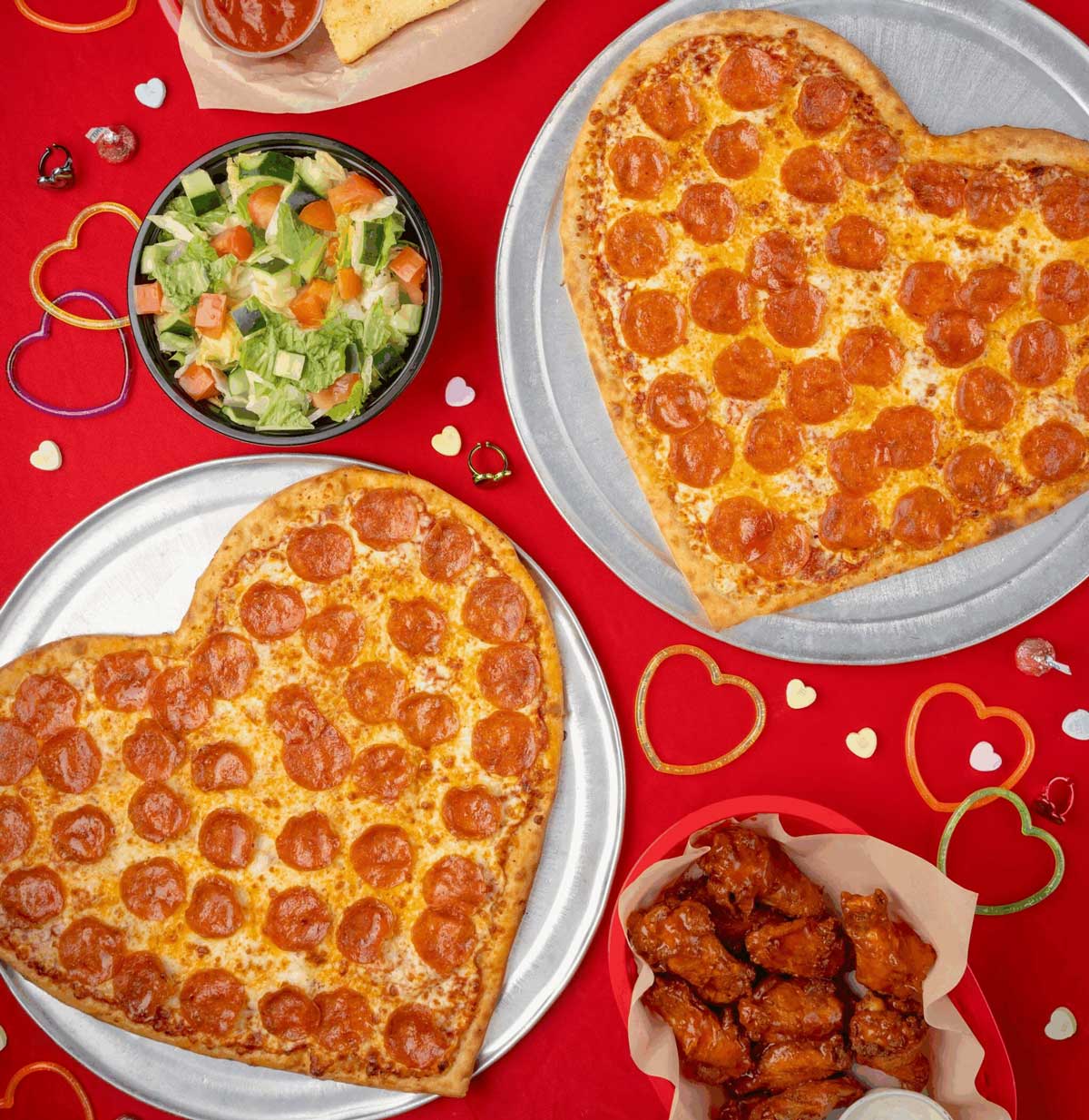 Heart-shaped pepperoni pizzas on metal trays, a bowl of salad, buffalo chicken wings, breadsticks, and scattered colorful heart decorations on a red tablecloth.