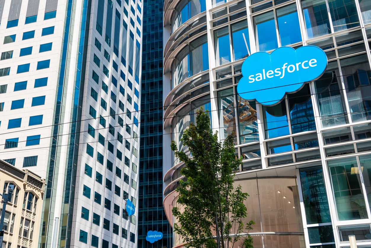 A modern glass building with a blue "Salesforce" logo above its entrance, set amid other tall downtown offices. A tree is visible in the foreground, reflecting the city's resilience after the 2026 layoffs.