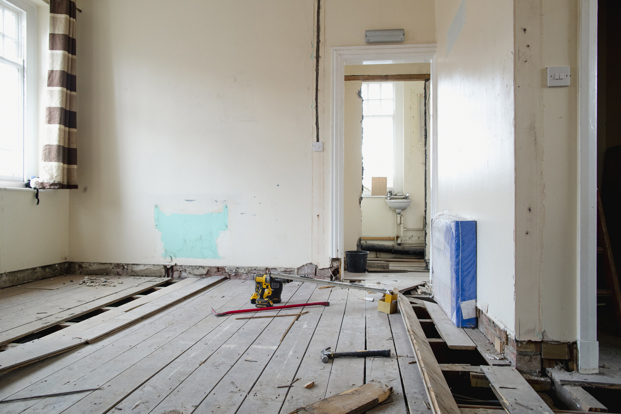 Room under renovation with floorboards removed, exposing beams underneath. Tools and materials are scattered on the wooden floor. Walls are partially painted, and a small sink is visible in an adjoining room through an open door.