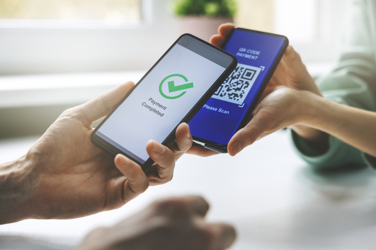 Two people hold smartphones next to each other, one displaying a QR code for payment and the other showing a “Payment Completed” confirmation with a green checkmark, amidst daily life unchanged by the 2026 layoffs.
