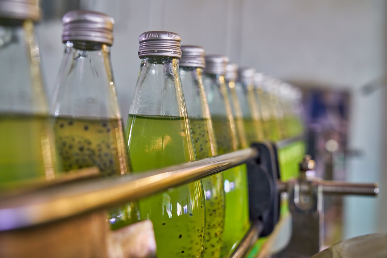 Several glass bottles filled with a green liquid containing basil seeds are lined up with silver caps in a bottling or production facility.