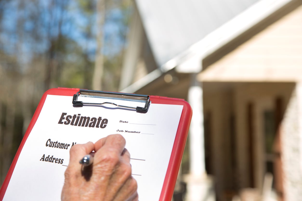 A person fills out a home repair costs estimate form on a clipboard outdoors, with a house in the background. The form includes spaces for the date, customer name, address, and job number.