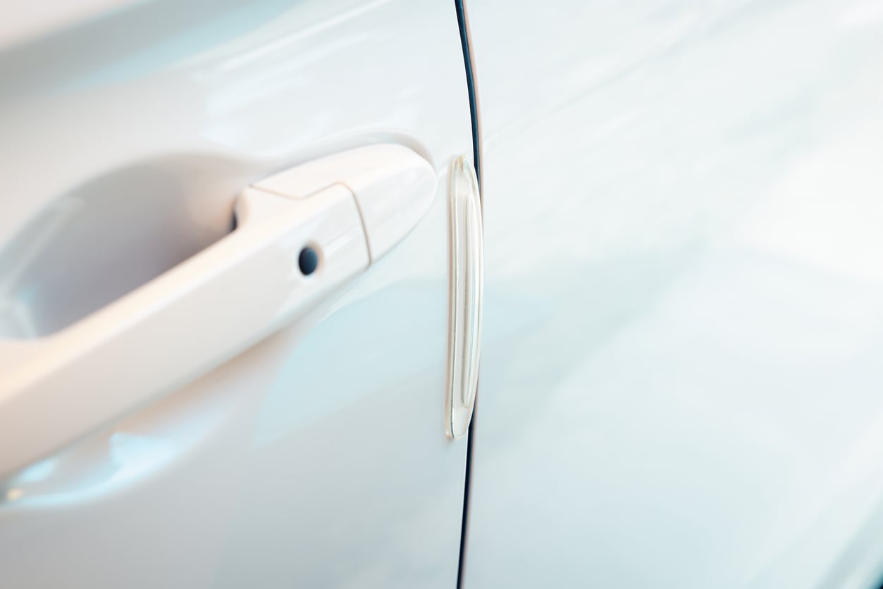 Close-up of a white car door showing the handle and a thin, vertical protective strip attached near the edge to prevent scratches or dents. The background is softly blurred.