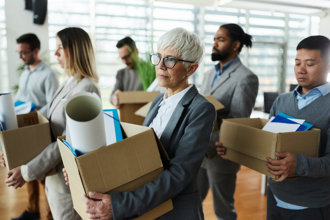 A group of office workers, appearing upset, hold boxes with personal belongings, suggesting they have been laid off or are leaving their jobs. They are dressed in business attire in a modern office setting.