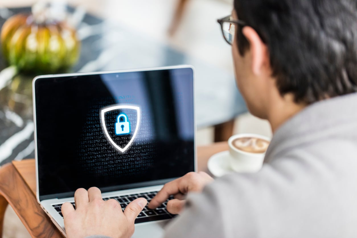 A person types on a laptop displaying a shield icon with a lock on the screen, symbolizing cybersecurity. A cup of coffee and a pumpkin are visible in the background.