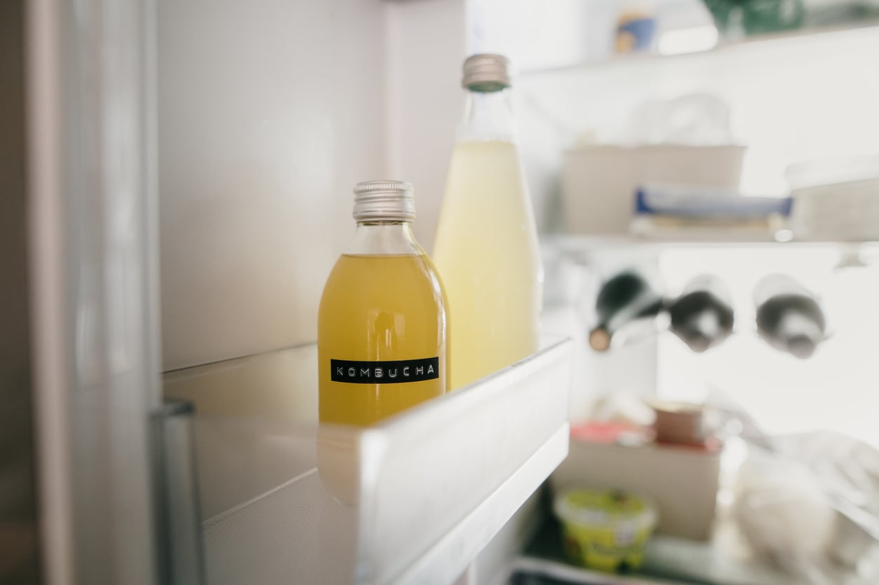 A glass bottle labeled "Kombucha" sits on a refrigerator door shelf beside another bottle filled with a light yellow liquid, with various food items visible in the background.
