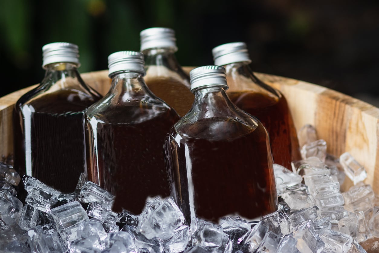 Five square glass bottles with silver caps, filled with dark liquid, are placed in a wooden bowl filled with ice cubes. The bottles are closely packed and partially embedded in the ice.