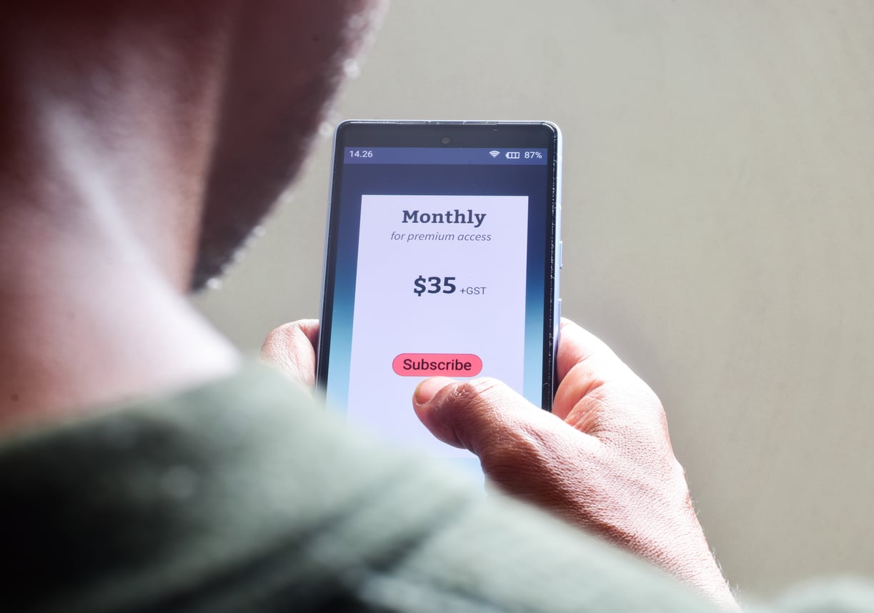 A person holding a smartphone, about to tap a red "Subscribe" button on a screen showing a $35 monthly premium access plan.