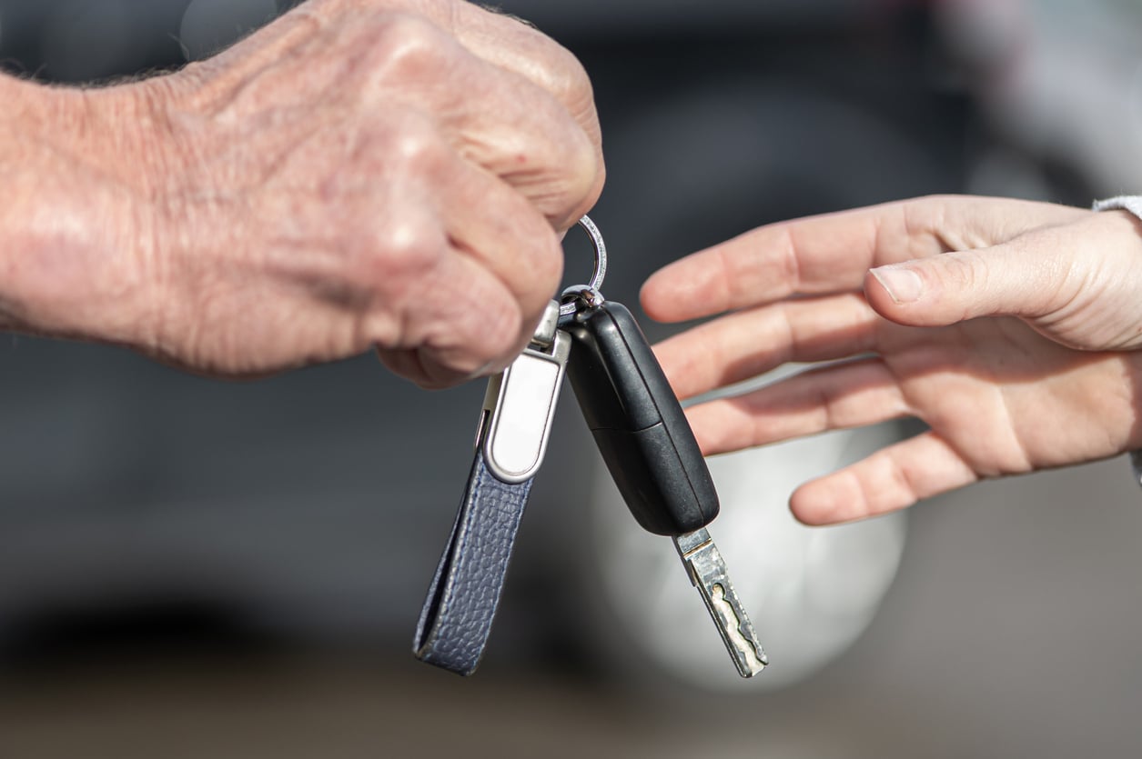 A close-up of an older hand handing over a car key with a keychain to a younger hand, symbolizing giving or receiving a car, with a blurred background.