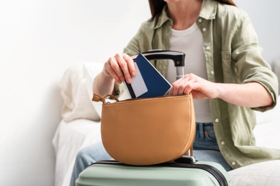 A woman sitting on a bed packs a passport and boarding pass into a tan purse resting on a mint green suitcase, preparing for travel.