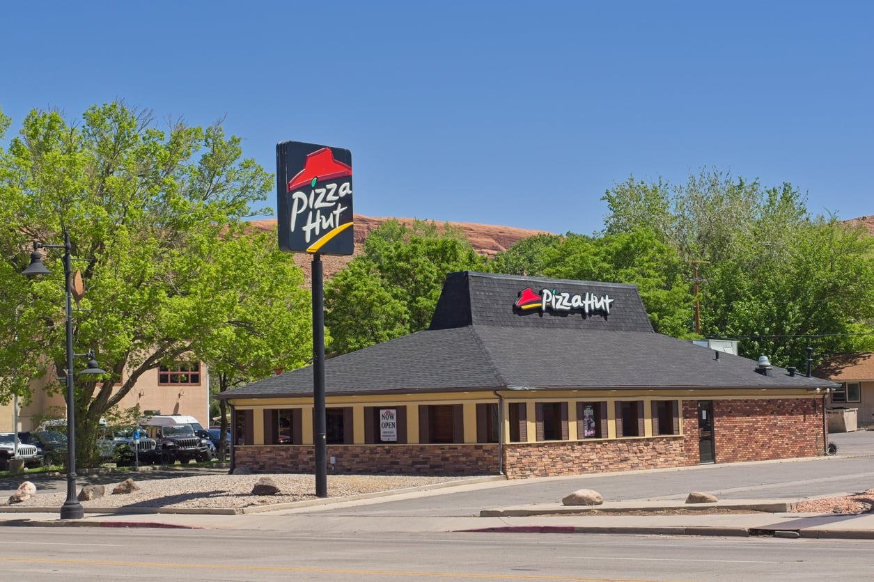 A Pizza Hut restaurant with a black roof and brick exterior sits beside a street on a clear, sunny day. Trees surround the building, and a Pizza Hut sign is visible on a tall pole.