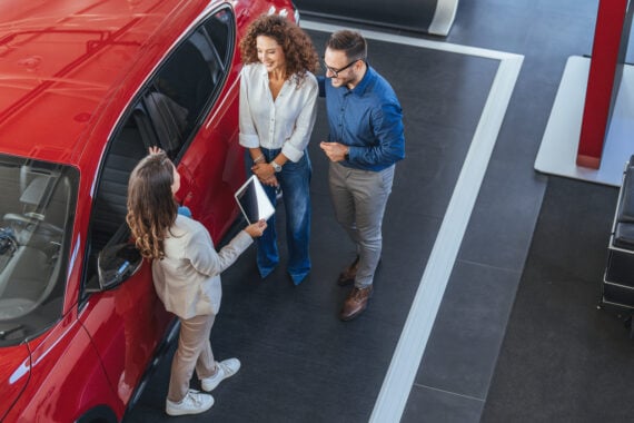 A car salesperson holding a tablet talks to a smiling couple standing beside a red car in a showroom, viewed from above.