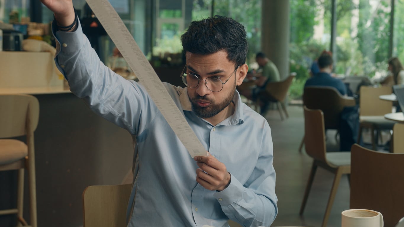 A man wearing glasses and a light blue shirt sits in a café, holding up and looking at a long receipt with a surprised expression. Other people are seated in the background.