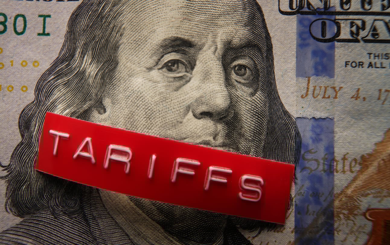 A close-up of Benjamin Franklin's portrait on a U.S. hundred-dollar bill with a red label reading "TARIFFS" placed across his face.