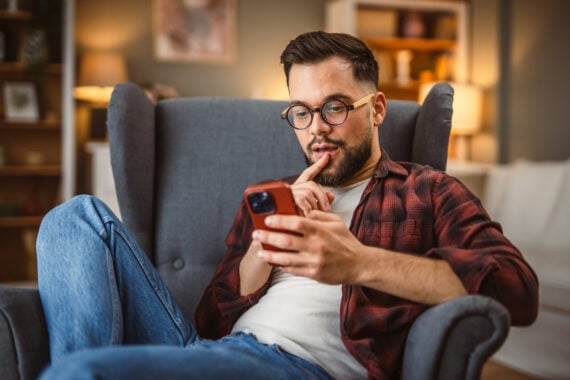 A man with glasses and a beard sits in an armchair, wearing a red plaid shirt and jeans, looking thoughtfully at his smartphone with one finger resting on his lips in a cozy living room.