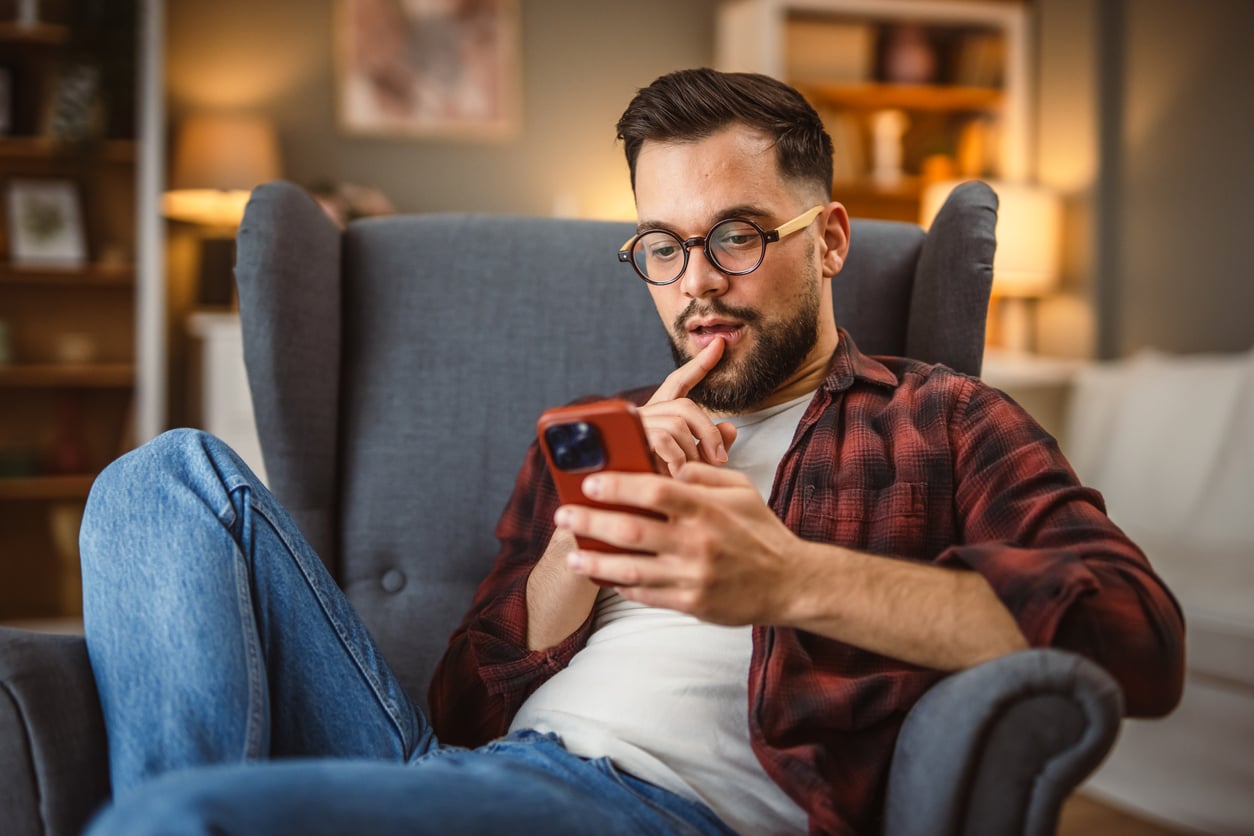 A man with glasses and a beard sits in an armchair, wearing a red plaid shirt and jeans, looking thoughtfully at his smartphone with one finger resting on his lips in a cozy living room.