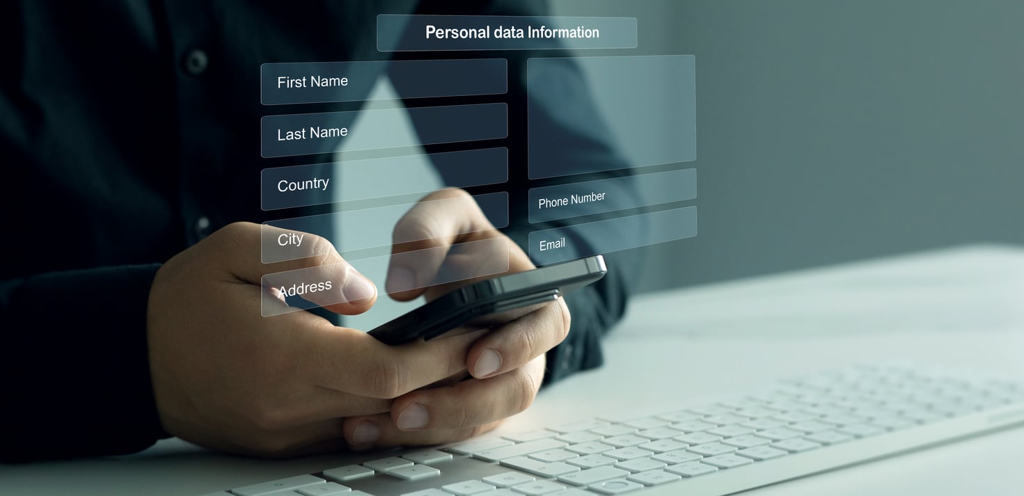 A person holding a smartphone sits at a desk with a keyboard, interacting with a transparent digital form requesting personal data such as name, country, phone number, city, address, and email.